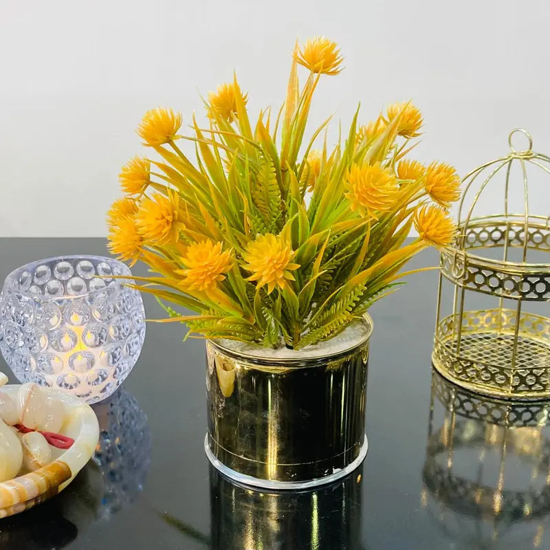Artificial yellow flowers in a pot on a reflective surface with decorative items.